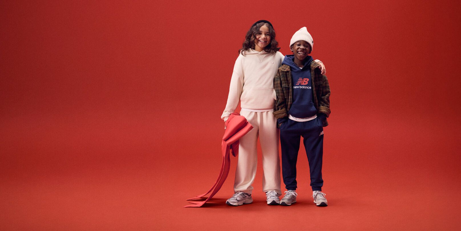 Two young kids smiling with arms around each other on a red background.