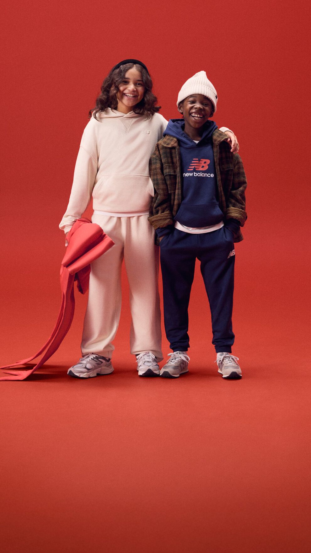 Two young kids smiling with arms around each other on a red background.